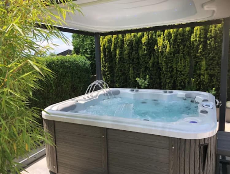 Freistehender Whirlpool mit Deckellift unter einer Pergola, dahinter eine Hecke
